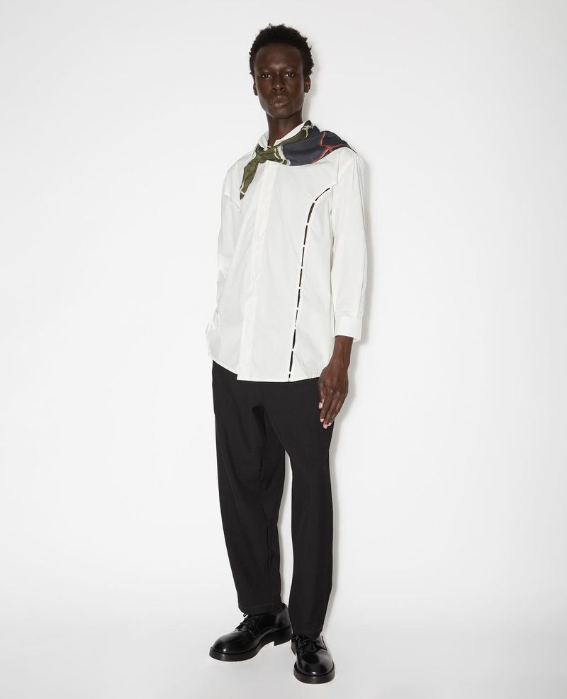 Man in a white shirt and black trousers, posing in a studio with a scarf around his neck, showcasing fashion style.