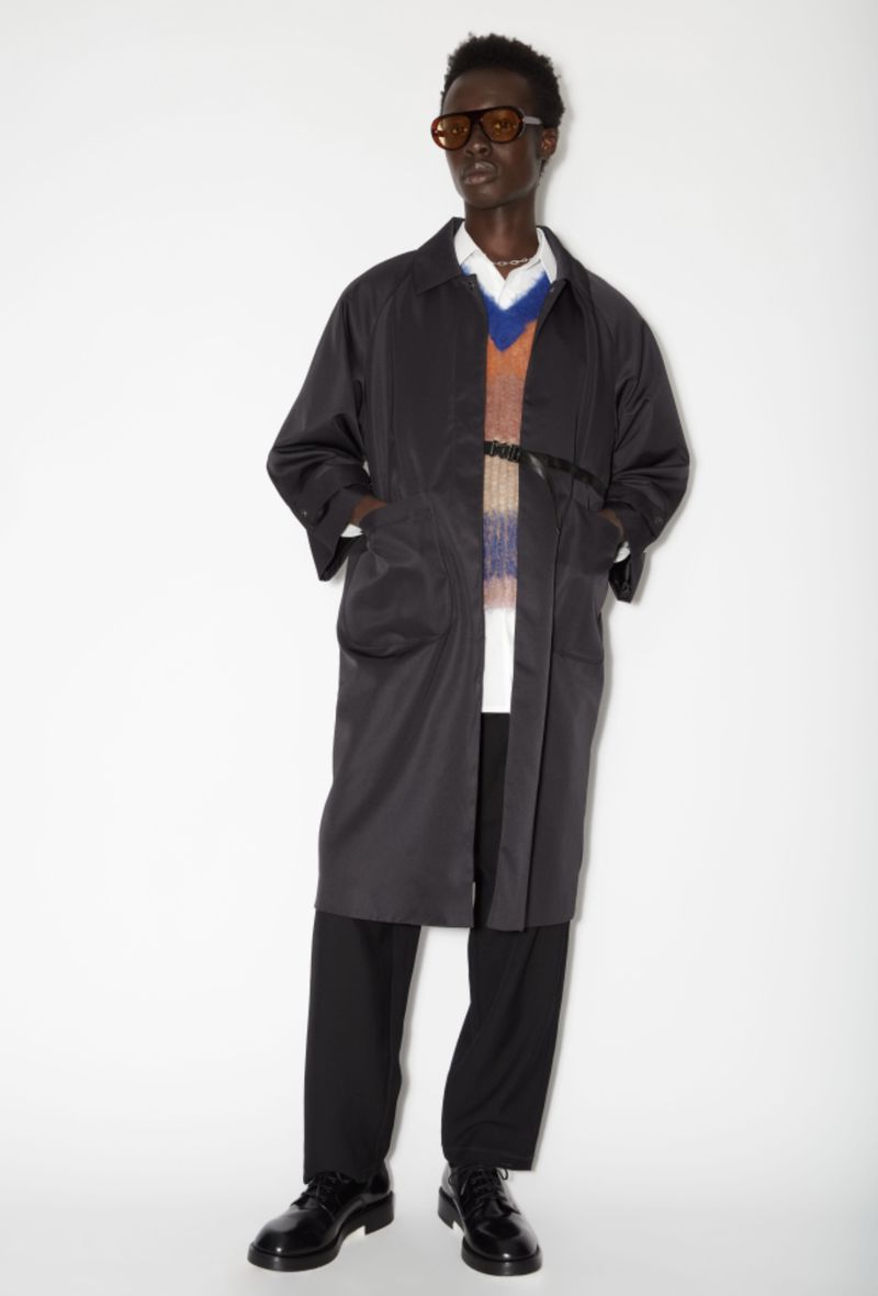 Mani Adjaye wearing a long dark overcoat, colorful knit sweater, and tinted sunglasses against a white studio backdrop.