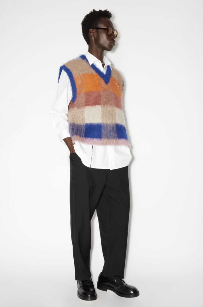 Man in a colorful checkered sweater vest, black pants, and black dress shoes poses in a full shot against a white backdrop.