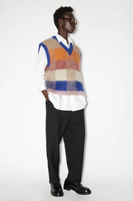 Man in a colorful checkered sweater vest, black pants, and black dress shoes poses in a full shot against a white backdrop.