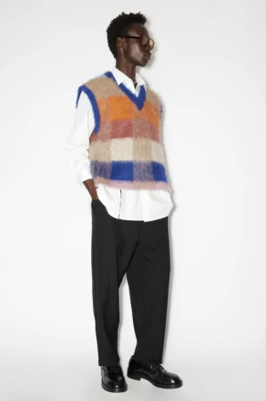 Man in a colorful checkered sweater vest, black pants, and black dress shoes poses in a full shot against a white backdrop.