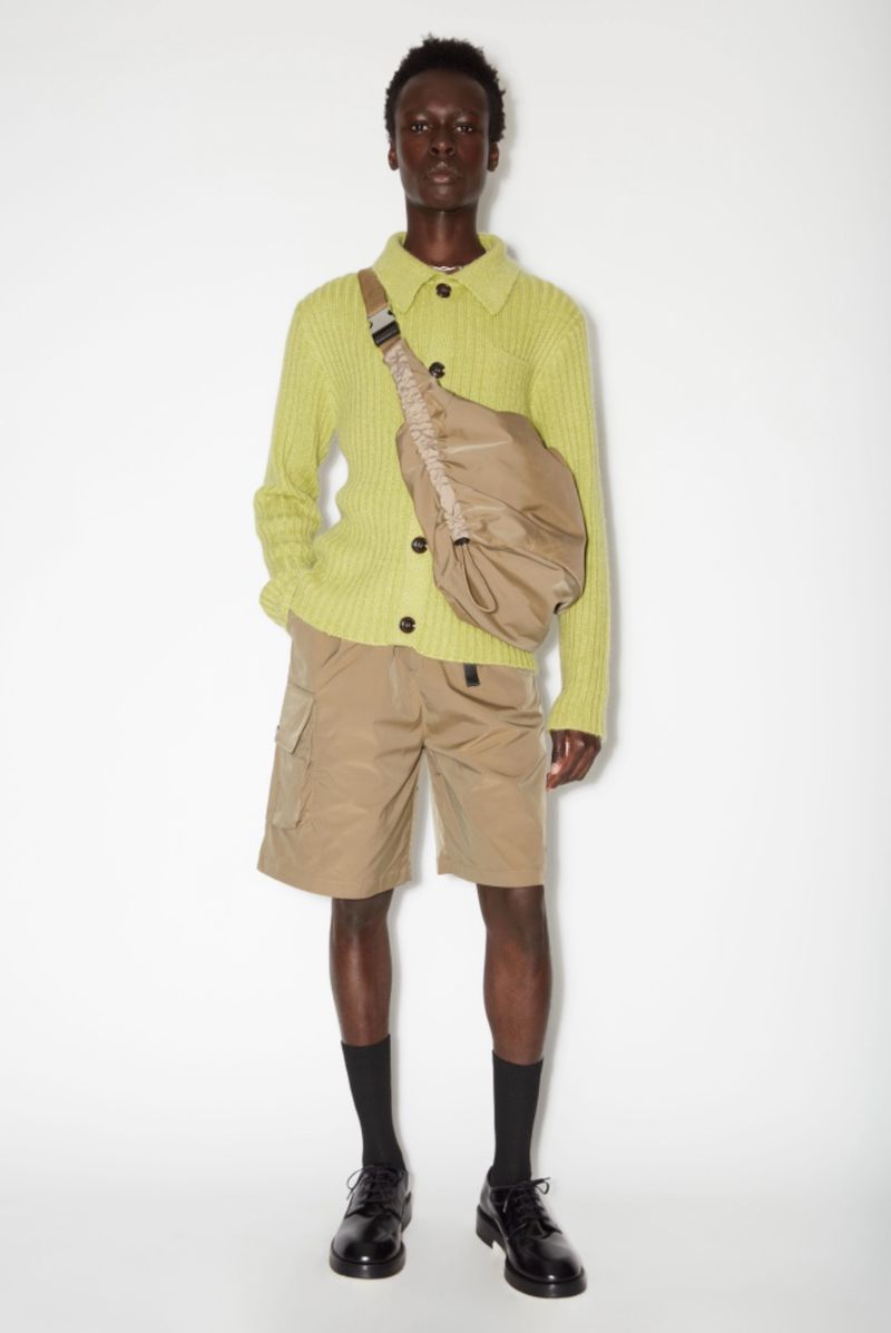 Male fashion model poses in a studio wearing a chartreuse sweater, khaki shorts, black socks, and dress shoes with a crossbody bag.