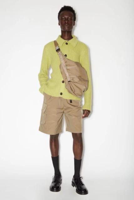 Male fashion model poses in a studio wearing a chartreuse sweater, khaki shorts, black socks, and dress shoes with a crossbody bag.
