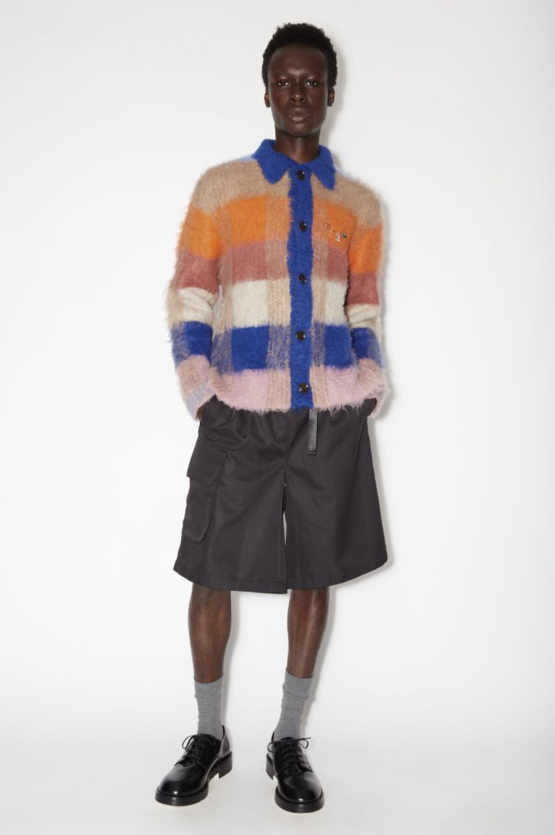 Full shot of a slender adult man in a fuzzy, multicolored, patterned shirt, black shorts, gray socks, and black dress shoes in a white studio.