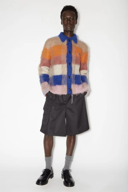 Full shot of a slender adult man in a fuzzy, multicolored, patterned shirt, black shorts, gray socks, and black dress shoes in a white studio.
