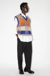Model wearing a fuzzy plaid sweater vest over a white shirt and dark trousers against a plain white background