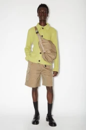 Model wearing a chartreuse knit cardigan and khaki shorts with a beige crossbody bag against a plain white background.