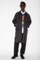 Mani Adjaye wearing a long dark overcoat, colorful knit sweater, and tinted sunglasses against a white studio backdrop.