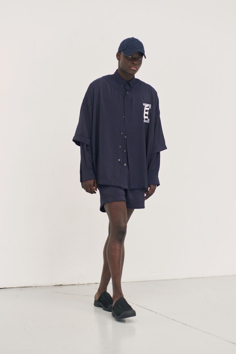A Black man in casual navy attire, including an oversized shirt, shorts, cap, and slides, poses in a well-lit, minimalist white studio.