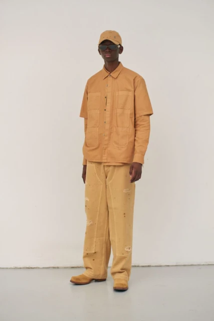 Full-length studio shot of a dark-skinned male model wearing a tan workwear-inspired outfit with jacket, jeans, baseball cap, and sunglasses.