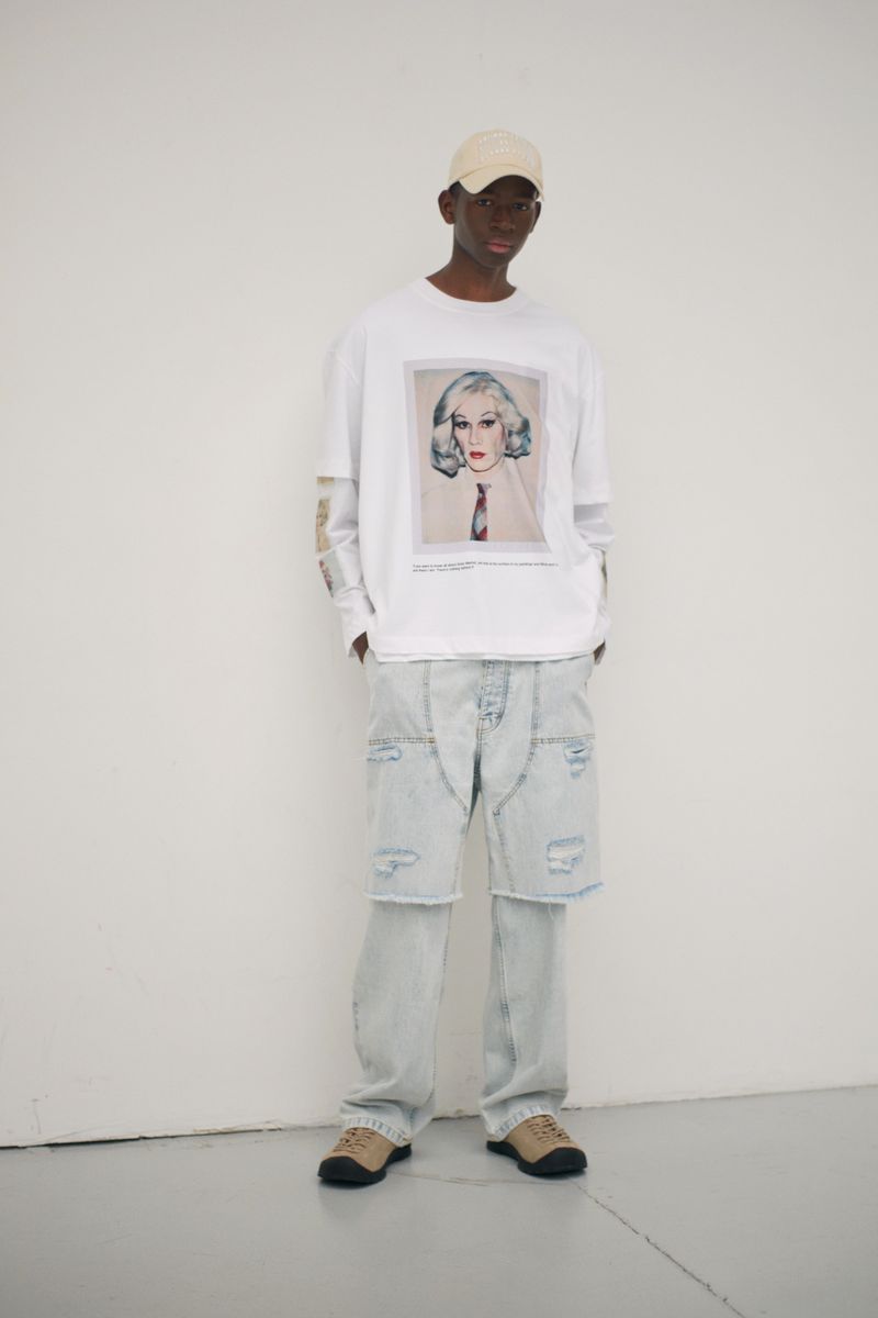 Full-body studio shot of a Black male model in a white graphic tee, deconstructed jeans, beige cap and sneakers against a white backdrop.