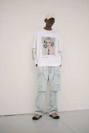 Full-body studio shot of a Black male model in a white graphic tee, deconstructed jeans, beige cap and sneakers against a white backdrop.