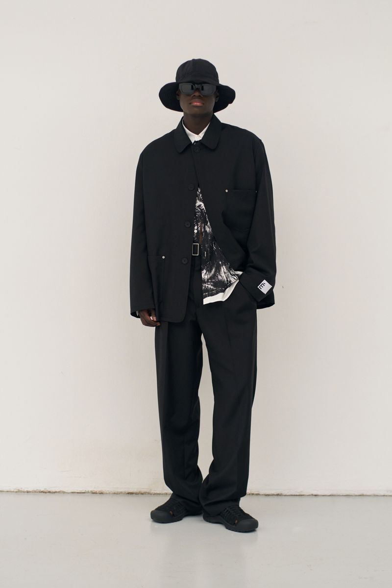 Full-length studio shot of a male fashion model in a black jacket and trousers, wearing a bucket hat and sunglasses against a neutral background.