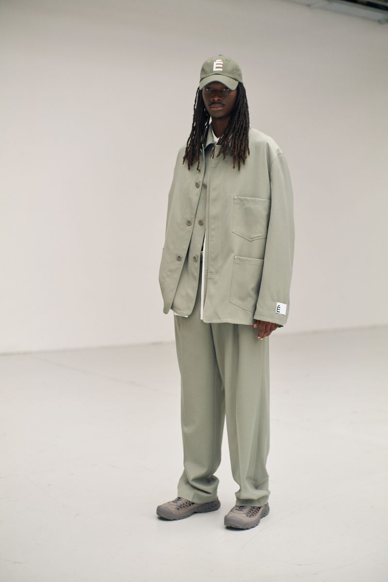 A person with long dreadlocks, wearing a light green jacket and pants with gray sneakers, standing in a white photo studio.