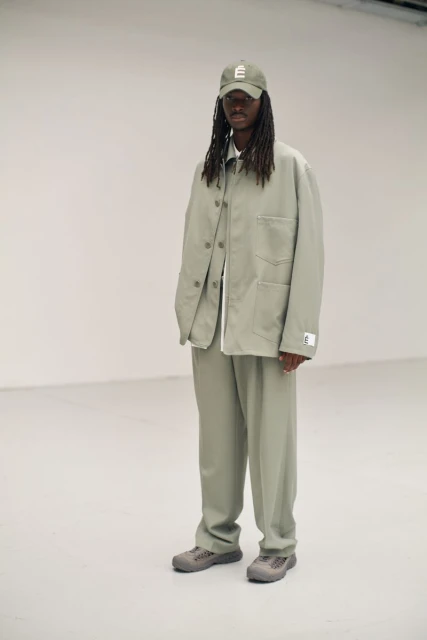 A person with long dreadlocks, wearing a light green jacket and pants with gray sneakers, standing in a white photo studio.