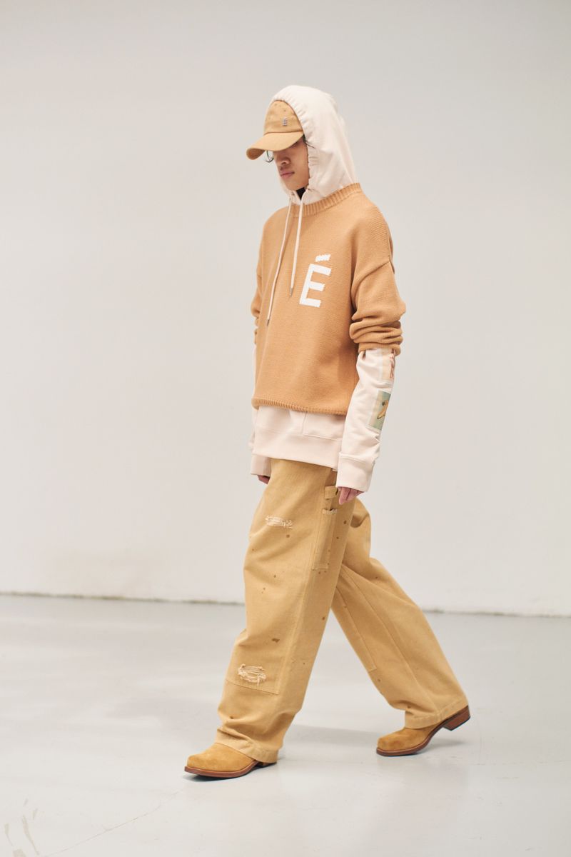 An adult male is walking in a photo studio. He is wearing a tan hoodie with an emblem on the front, loose tan pants, brown shoes, and a baseball cap.