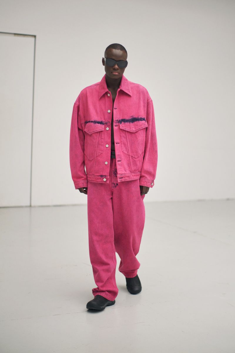 An adult Black male in a pink denim jacket and matching pants poses in a minimalist studio with sunglasses and black shoes for a fashion editorial.