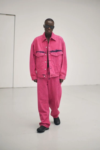 An adult Black male in a pink denim jacket and matching pants poses in a minimalist studio with sunglasses and black shoes for a fashion editorial.