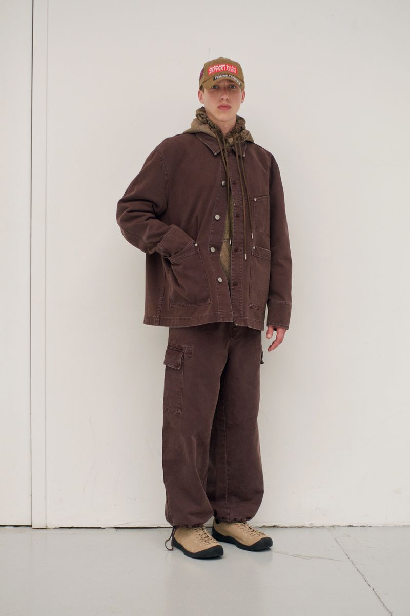 Full-length shot of an adult male in brown workwear outfit including jacket, pants, hoodie, baseball cap, and sneakers against a white wall.