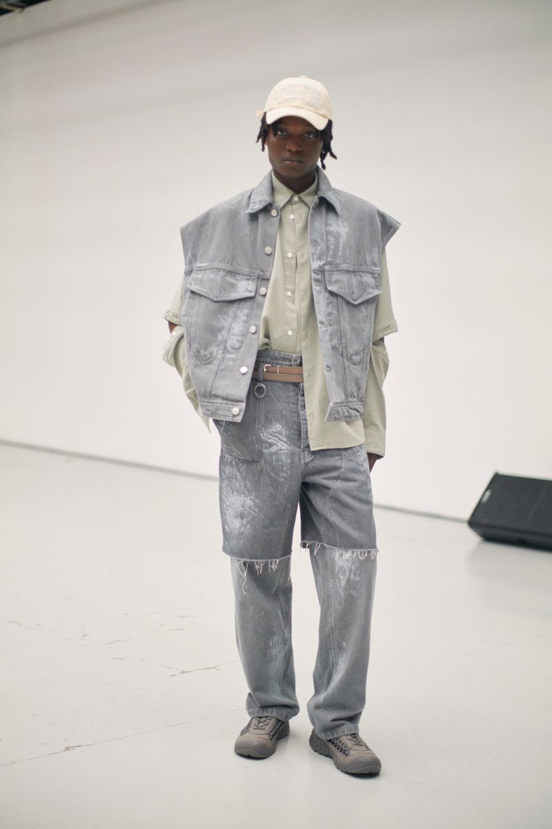 Full shot of an adult Black male model in a gray denim outfit with a cap, posing indoors against a plain white backdrop in a fashion editorial.