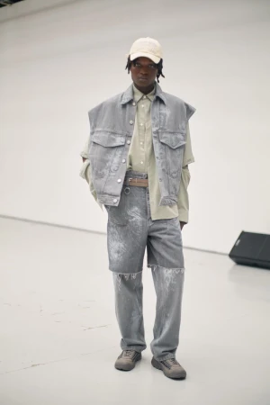 Full shot of an adult Black male model in a gray denim outfit with a cap, posing indoors against a plain white backdrop in a fashion editorial.