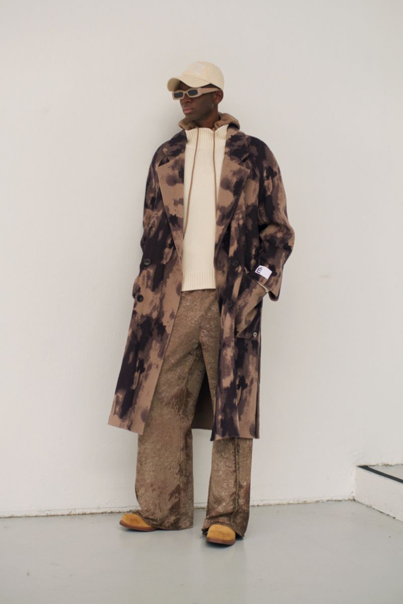 A Black male fashion model poses in a tie-dye patterned overcoat, hoodie, and wide-leg trousers against a neutral wall in a studio shot.