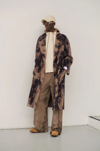 A Black male fashion model poses in a tie-dye patterned overcoat, hoodie, and wide-leg trousers against a neutral wall in a studio shot.