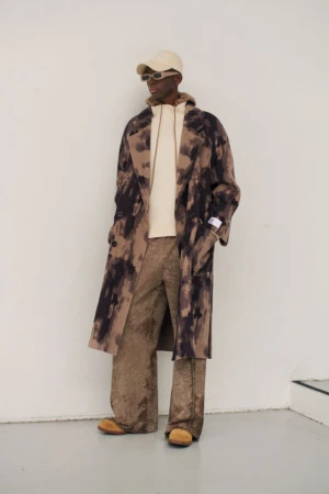 A Black male fashion model poses in a tie-dye patterned overcoat, hoodie, and wide-leg trousers against a neutral wall in a studio shot.