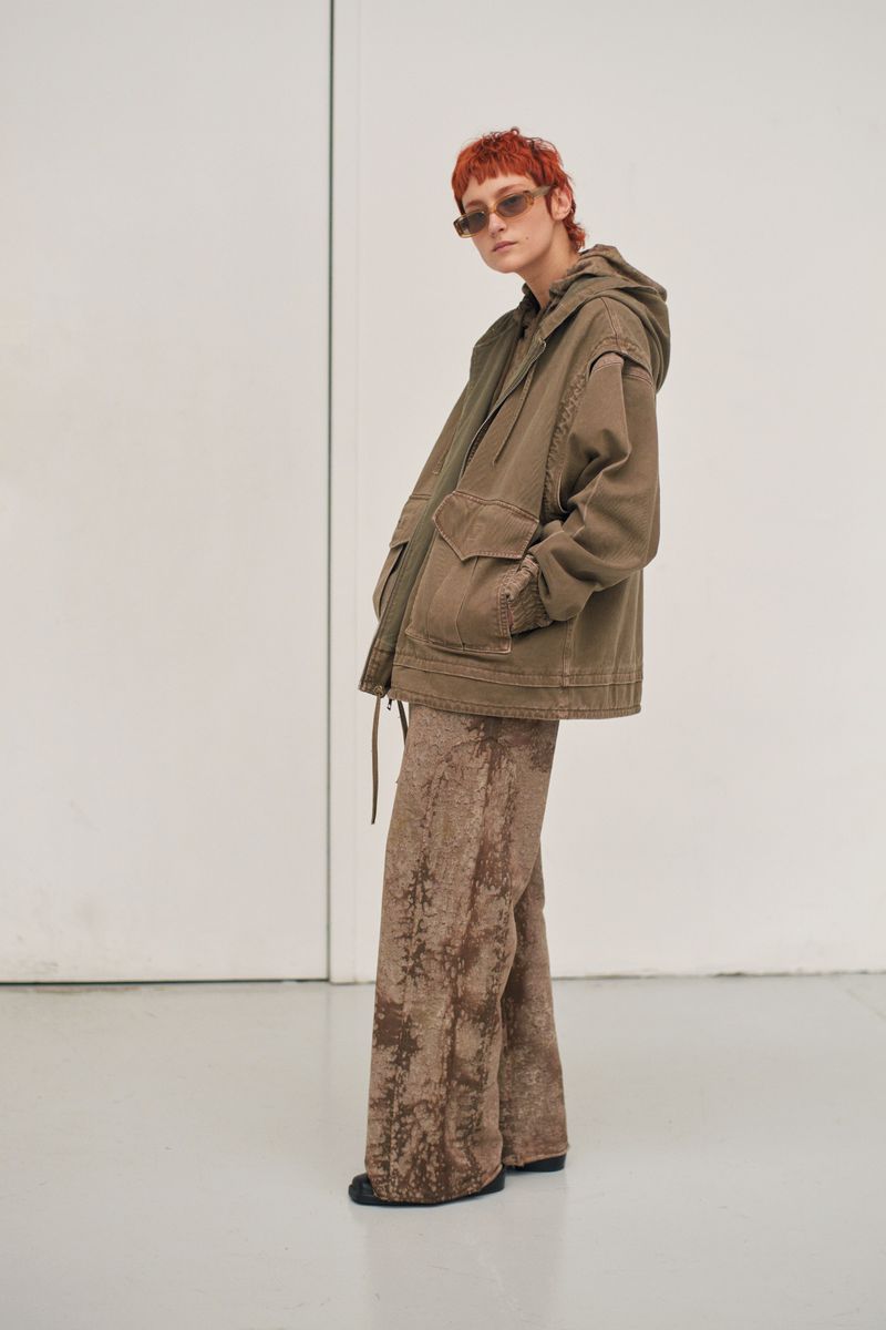 Fashion portrait of a model wearing a hooded olive green jacket and wide-leg textured pants standing in front of a white wall.
