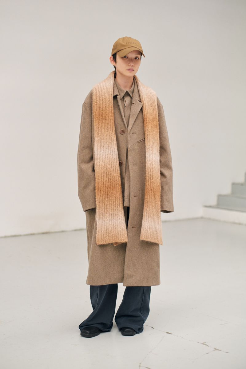 A person wearing a coat, scarf, and cap poses in a minimalist indoor setting with neutral tones and natural light, showcasing a fashion aesthetic.