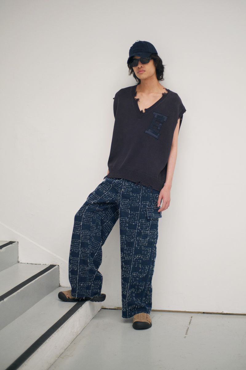 Young adult man in a casual outfit with a sleeveless shirt, patterned pants, baseball cap, and sunglasses standing on stairs against white wall.