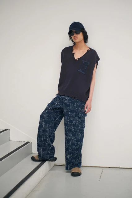 Young adult man in a casual outfit with a sleeveless shirt, patterned pants, baseball cap, and sunglasses standing on stairs against white wall.