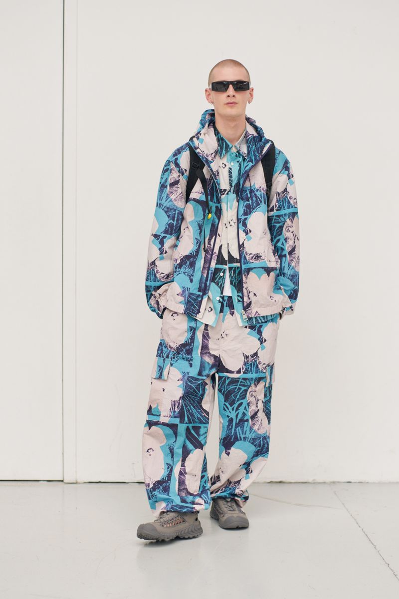 Male model poses in an indoor photo studio, showcasing a blue and white patterned jacket, matching pants, and sunglasses for fashion editorial.