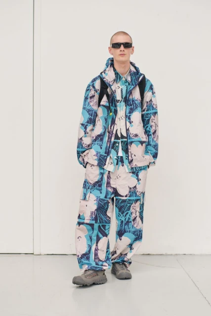 Male model poses in an indoor photo studio, showcasing a blue and white patterned jacket, matching pants, and sunglasses for fashion editorial.