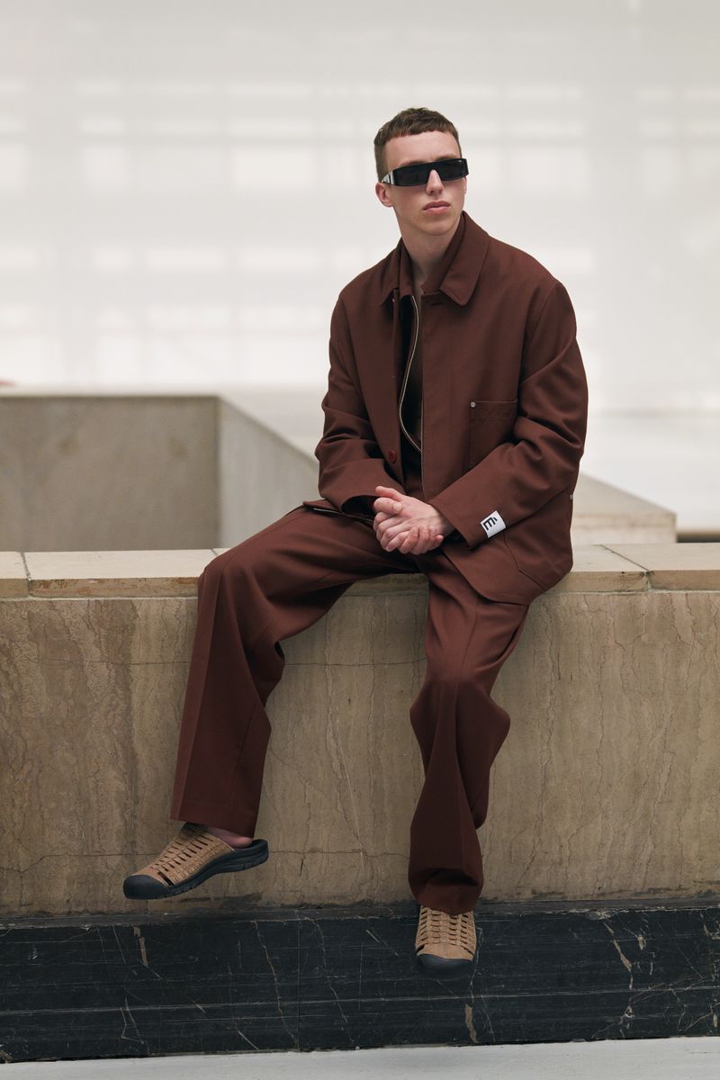 An adult man sits on a stone ledge in a brown suit and dark sunglasses in a brightly lit indoor space, showcasing a fashion editorial look.