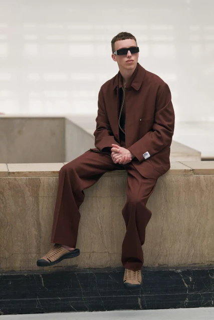 An adult man sits on a stone ledge in a brown suit and dark sunglasses in a brightly lit indoor space, showcasing a fashion editorial look.