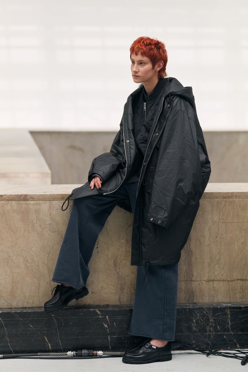 Woman with short auburn hair in a black hooded raincoat and blue pants, seated, posing in a neutral indoor environment.