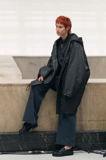 Woman with short auburn hair in a black hooded raincoat and blue pants, seated, posing in a neutral indoor environment.