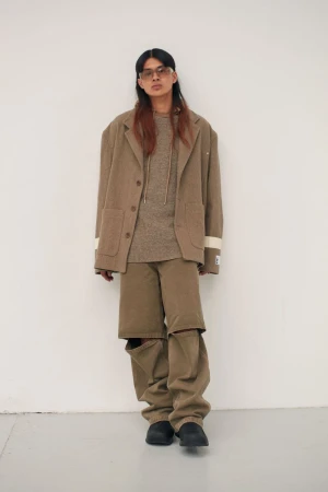 Male model wearing an oversized brown blazer, textured top, and cutout trousers with tinted sunglasses against a white background.