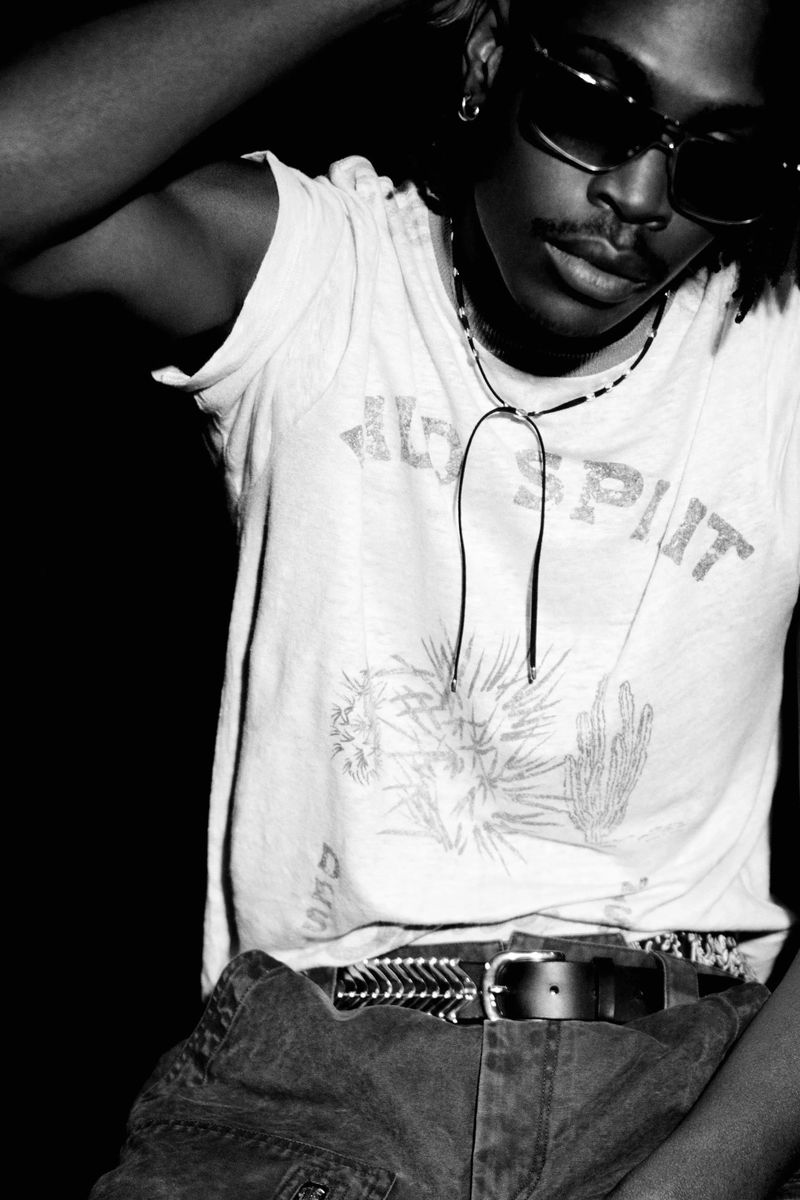 Monochrome portrait of an adult Black man wearing sunglasses, jeans, and graphic tee with a lasso bolo tie in a studio setting.
