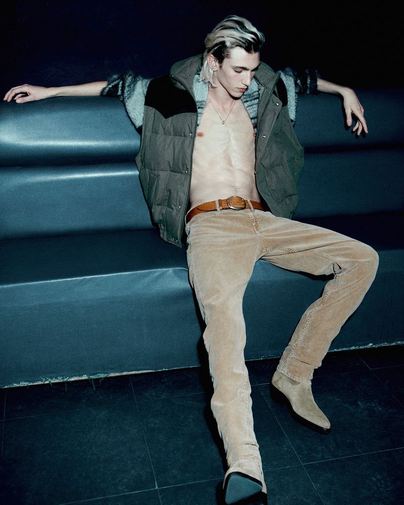 Stylish male model poses while sitting on a blue tiered bench, wearing a vest, pants, and boots in an indoor setting with dark lighting.