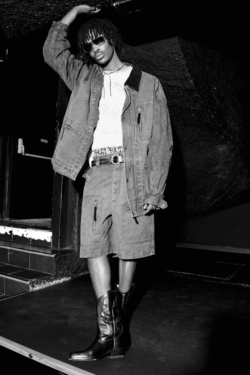 Monochrome portrait of a male model in a denim jacket, matching shorts, and tall leather boots with his hand touching the ceiling.