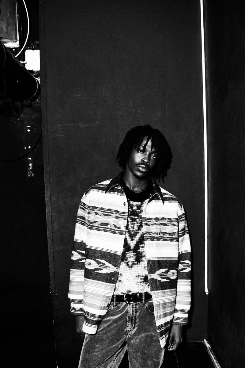 A young adult man poses in a patterned shirt and jeans in a studio with dark walls and soft lighting, creating a stylish fashion image.