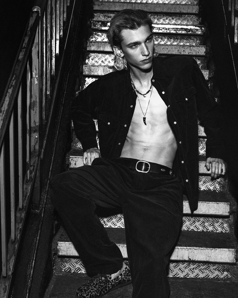 Black and white portrait of a young adult male with bare torso posing on metal stairs wearing black velvet suit and animal print shoes.
