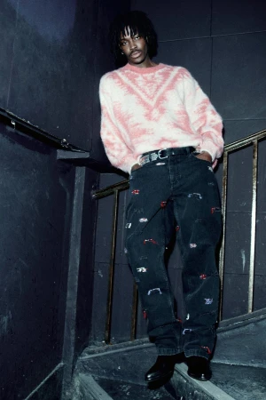 An adult male with locs, wearing a light pink sweater and decorated jeans, poses on stairs with a dark background.