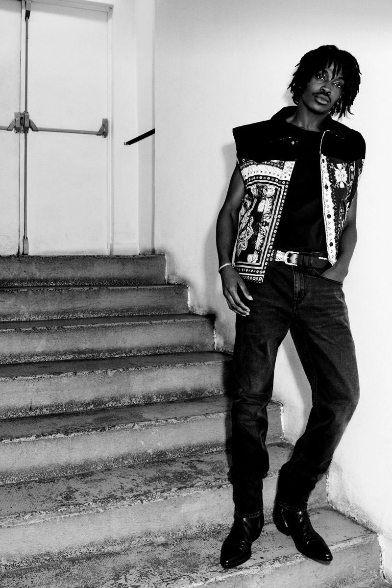 An adult male with dark hair, a sleeveless top, and jeans poses on concrete steps in a black and white, eye-level medium shot.