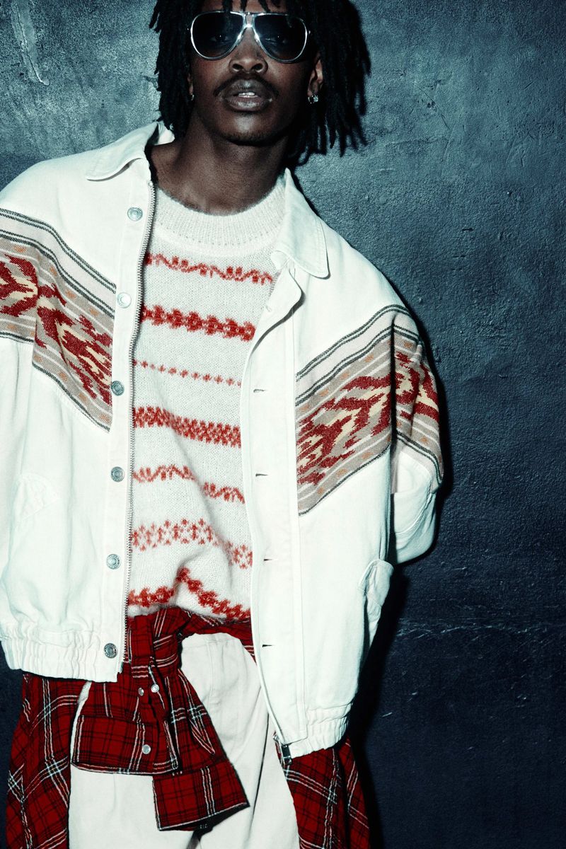 Man with dreadlocks wears aviator sunglasses, patterned sweater, and white jacket against a textured dark blue wall in a medium shot.