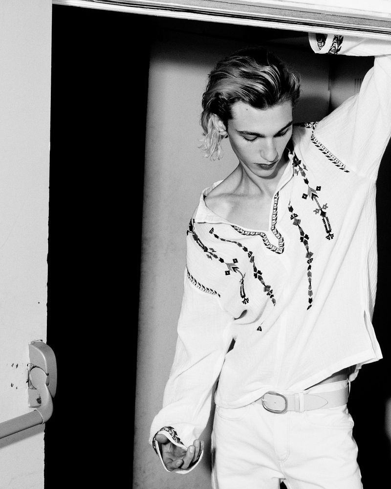 Black and white portrait of a slender, blonde adult male wearing a white, patterned shirt and white jeans in an indoor doorway setting.