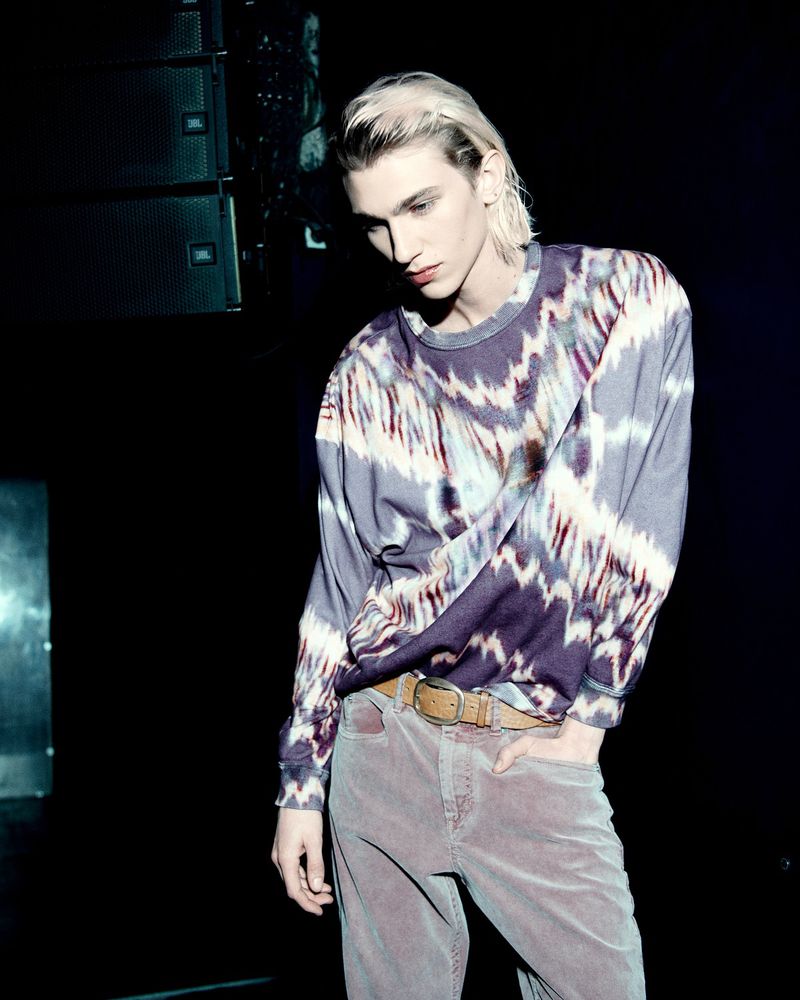 Portrait of a young adult male model in a patterned shirt and pants, posing indoors in front of JBL speakers, looking down.
