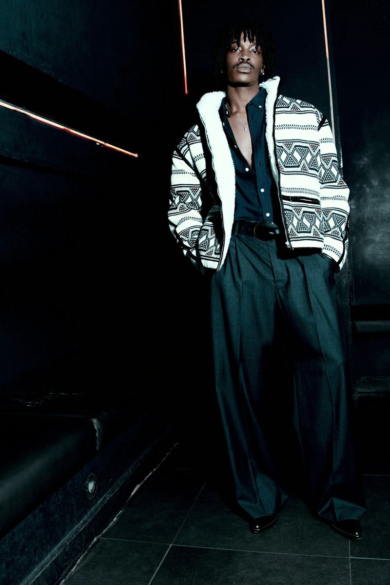Man in patterned jacket posing in dark indoor setting, wearing button-down shirt and wide-leg trousers. Fashion editorial photography.